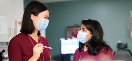 two females in maroon scrubs and masks talking in clinic
