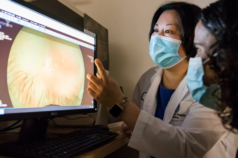 student looks on as faculty in white coat points at screen that is displaying eye imagining